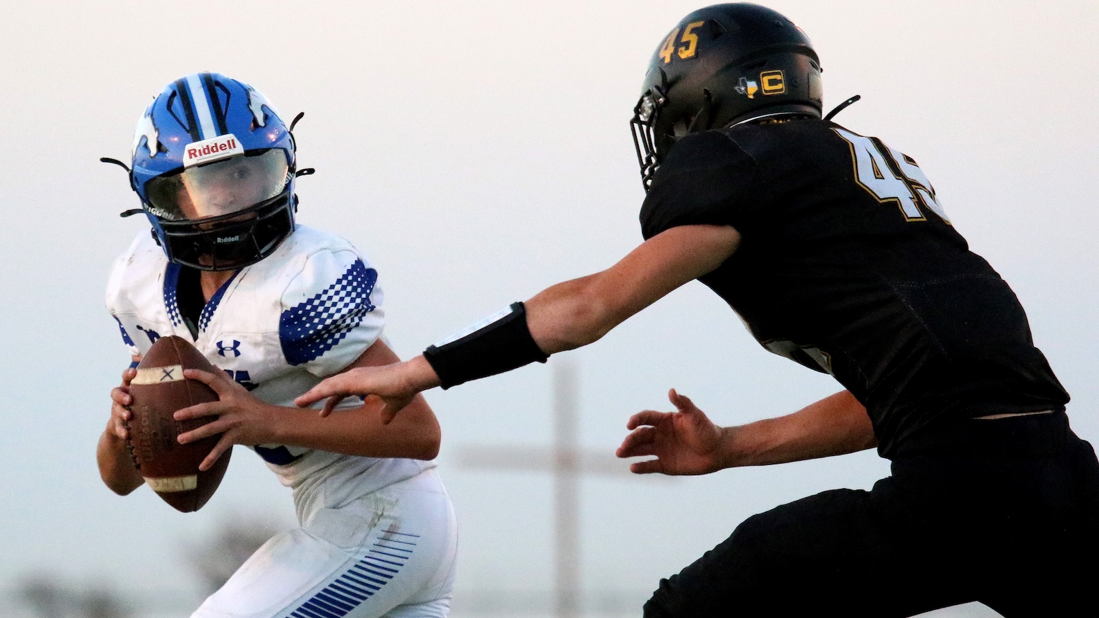 Quarterback in white and blue uniform holds the football while a defender in black jersey with number 45 reaches toward him in a tackle.