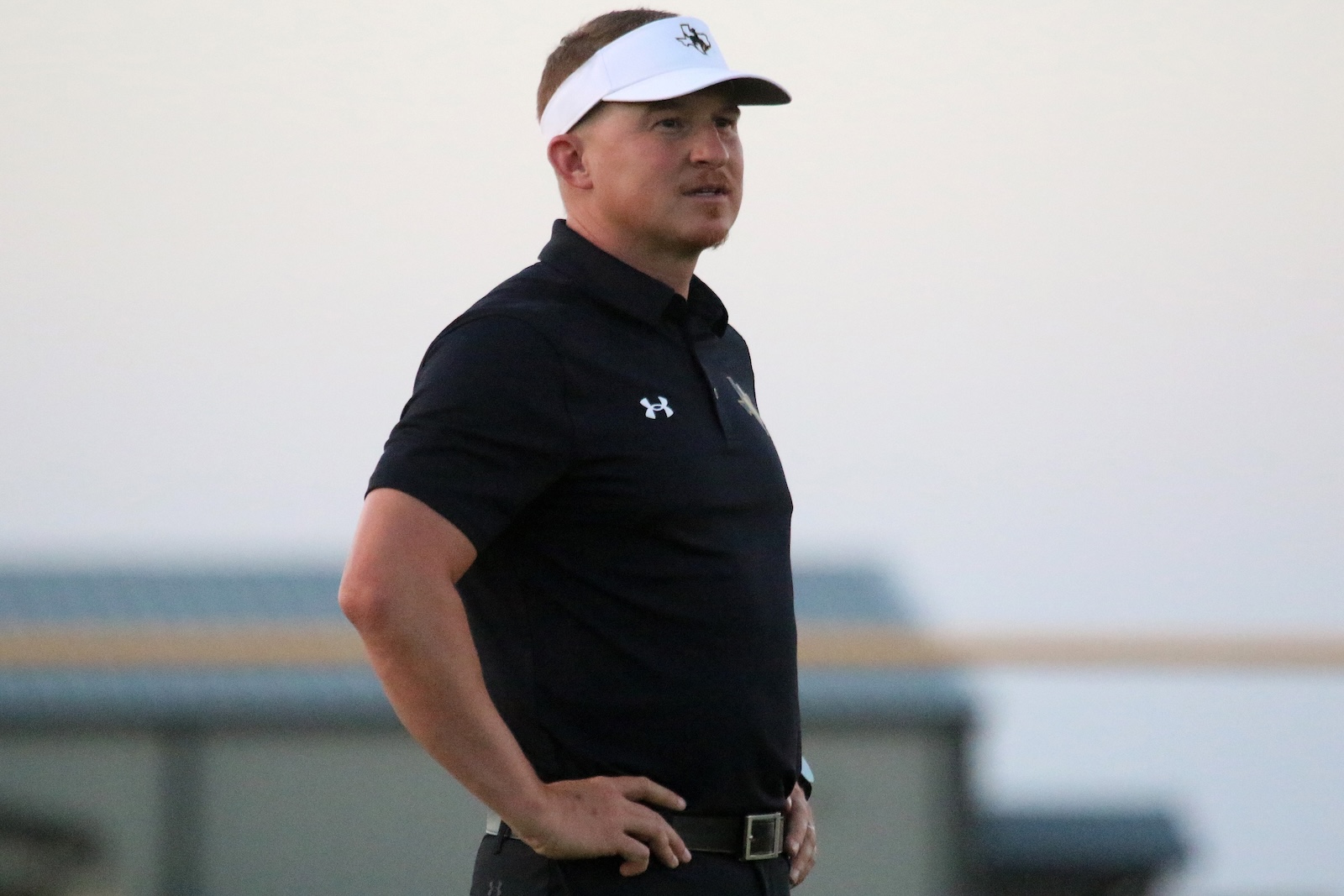 Man in a black polo with hands on hips wearing a white visor, standing on a sports field.