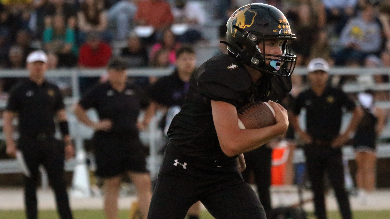 Football player in black uniform runs with the ball during a game, crowd blurred in the background, helmet on.