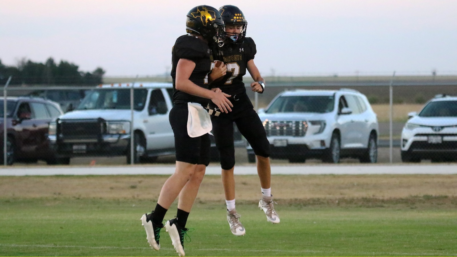 Two football players in black uniforms with gold helmets leap midair in celebration on a grassy field, with parked cars and a chain-link fence in the background.