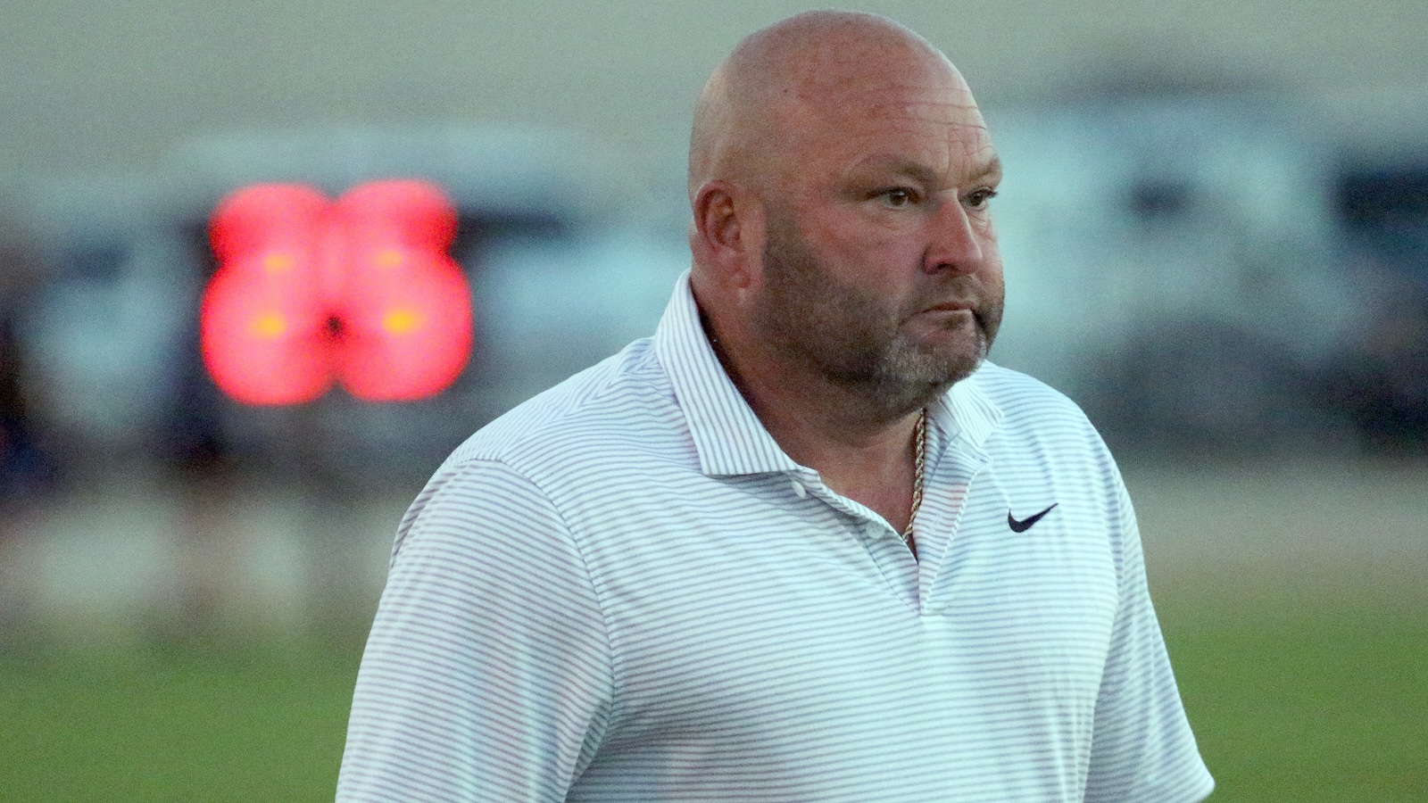 Bald man in a light striped polo shirt standing on a grassy sports field with a blurred red scoreboard in the background.