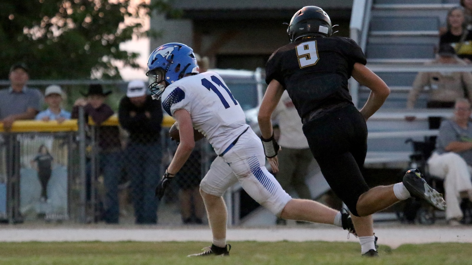 A football player in a white uniform with blue helmet runs while being chased by a player in a black uniform on a grassy field, with spectators in the background.
