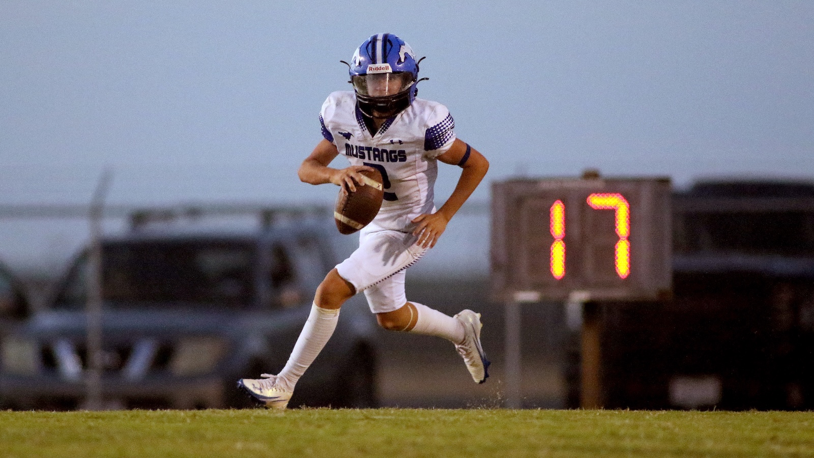 Football player in a white Mustangs uniform and blue helmet runs with the ball on a grassy field, a 17 on the scoreboard in the background.