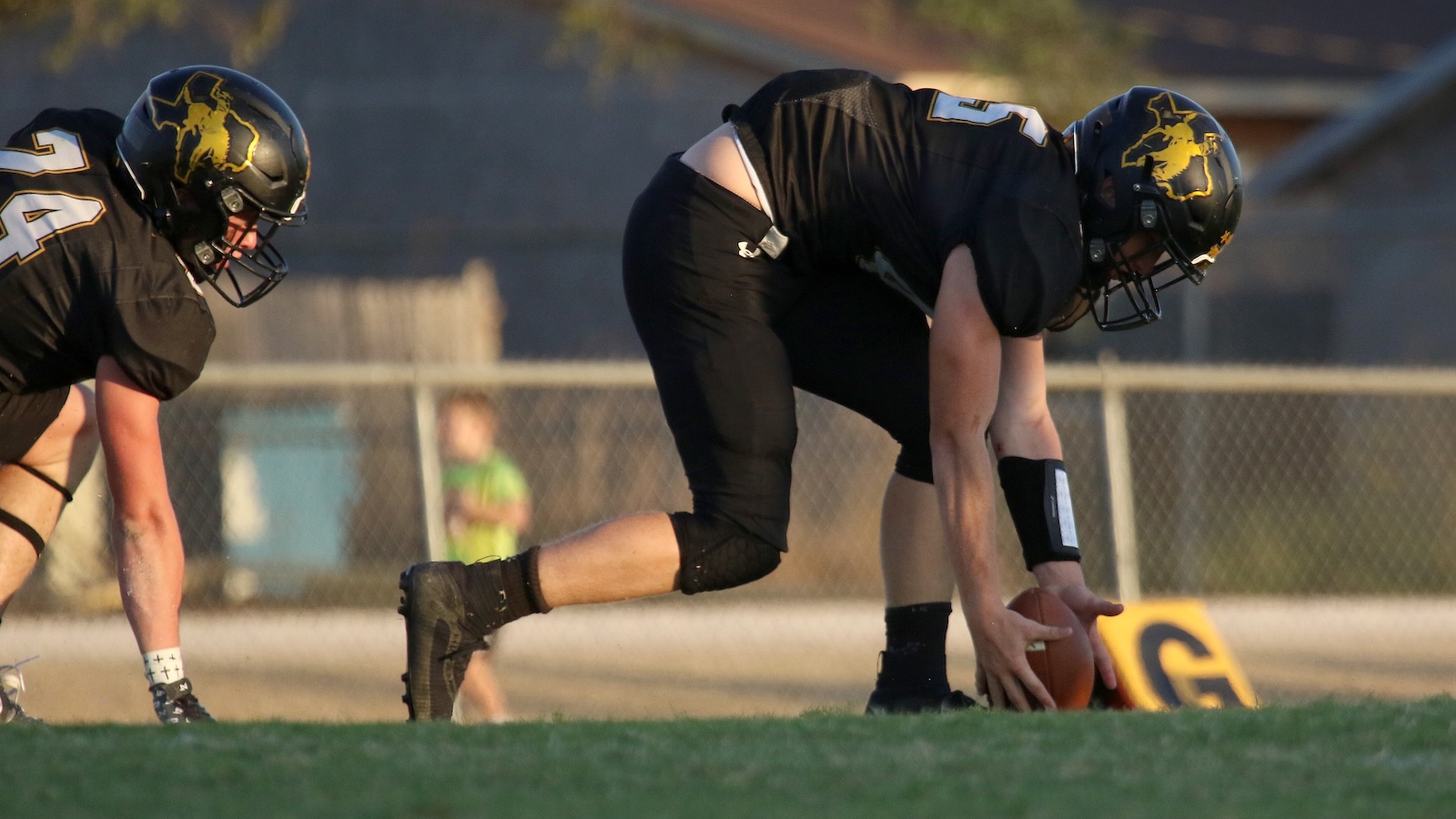Two football players in black uniforms line up at the snap; the center places the ball on the ground for the play.