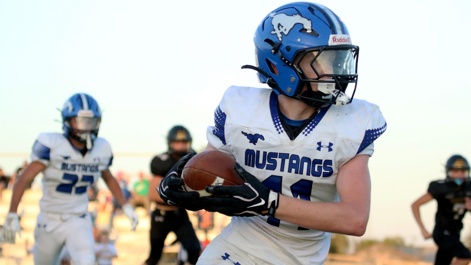 Boy in blue and white Mustang football uniform runs with the ball toward the camera, wearing a blue helmet with a mustang logo.