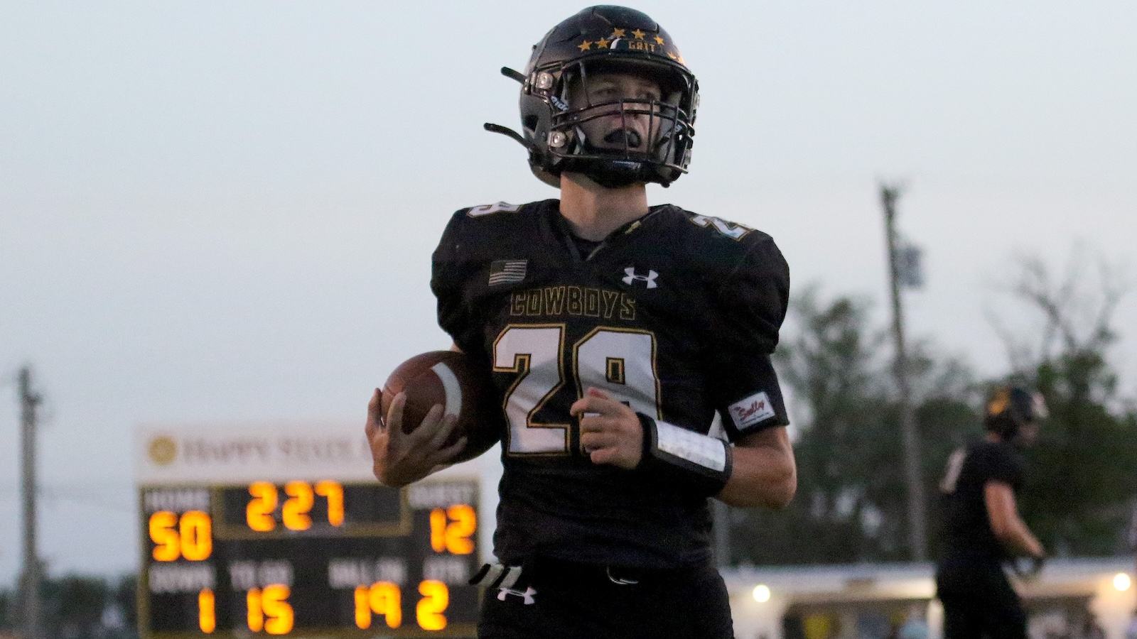 Football player in black Cowboys jersey #28 holding a ball and running on the field, scoreboard in the background.