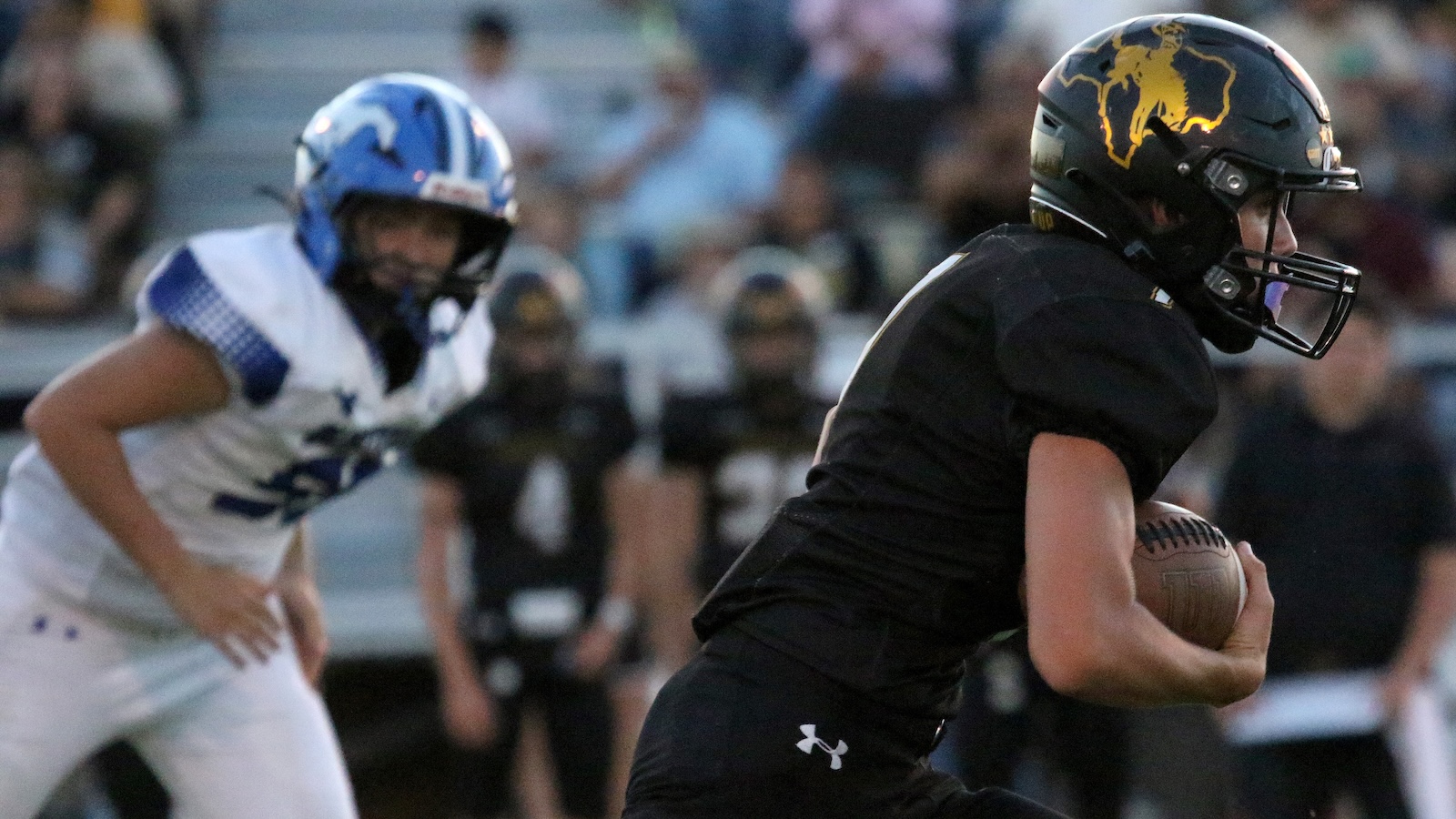 Football player in black uniform runs with ball during a game, a defender in white pursues in the background.