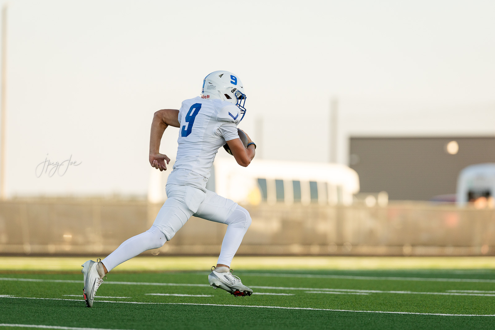 lubbock-christian-vs-bushland-football-jpegJoe-1 | Press Pass Sports American football player in a white uniform with the number 9 running with the ball on a green turf field under daylight, stadium backdrop in the distance
