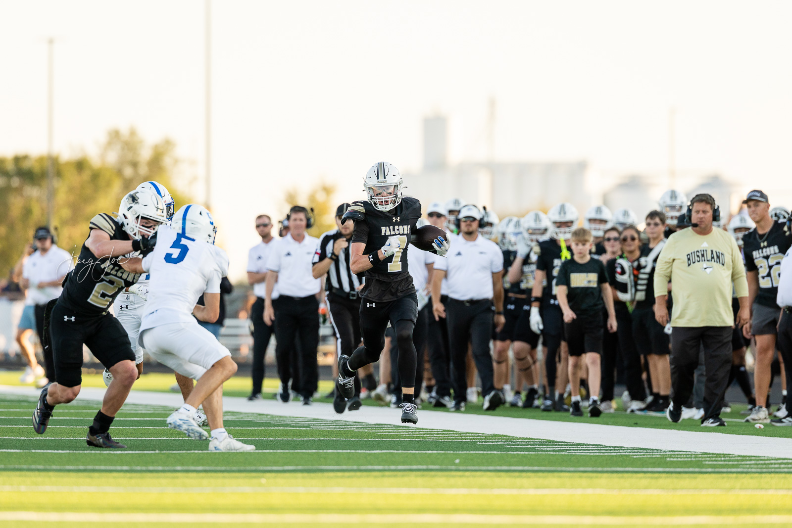 lubbock-christian-vs-bushland-football-jpegJoe-3 | Press Pass Sports Football game: a Black uniformed Falcons player with the ball runs upfield while white-uniformed defenders chase nearby on a bright turf field, with a crowd on the sideline.