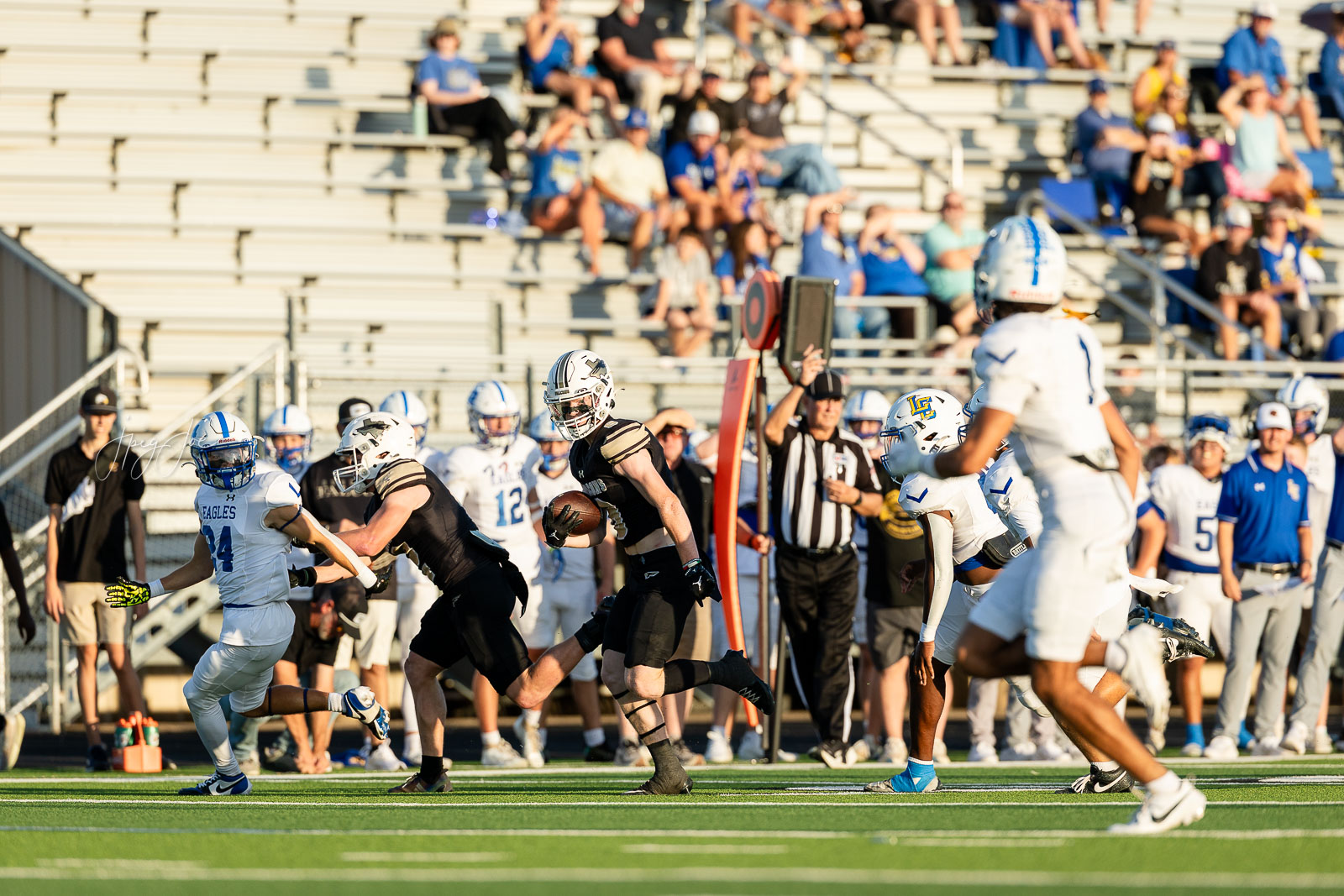lubbock-christian-vs-bushland-football-jpegJoe-4 | Press Pass Sports Football game in progress: a ball carrier in black runs with defenders close behind on a bright field, with fans in bleachers watching.
