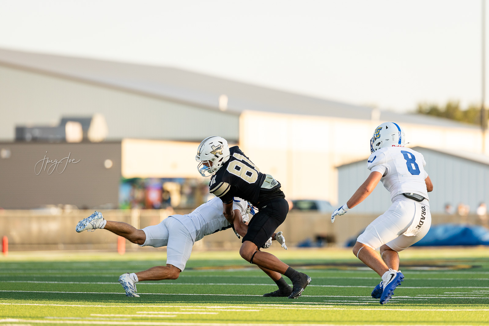 lubbock-christian-vs-bushland-football-jpegJoe-5 | Press Pass Sports A football player in a black jersey is tackled by a white-uniformed defender on a bright green field with a teammate in white watching in the background.