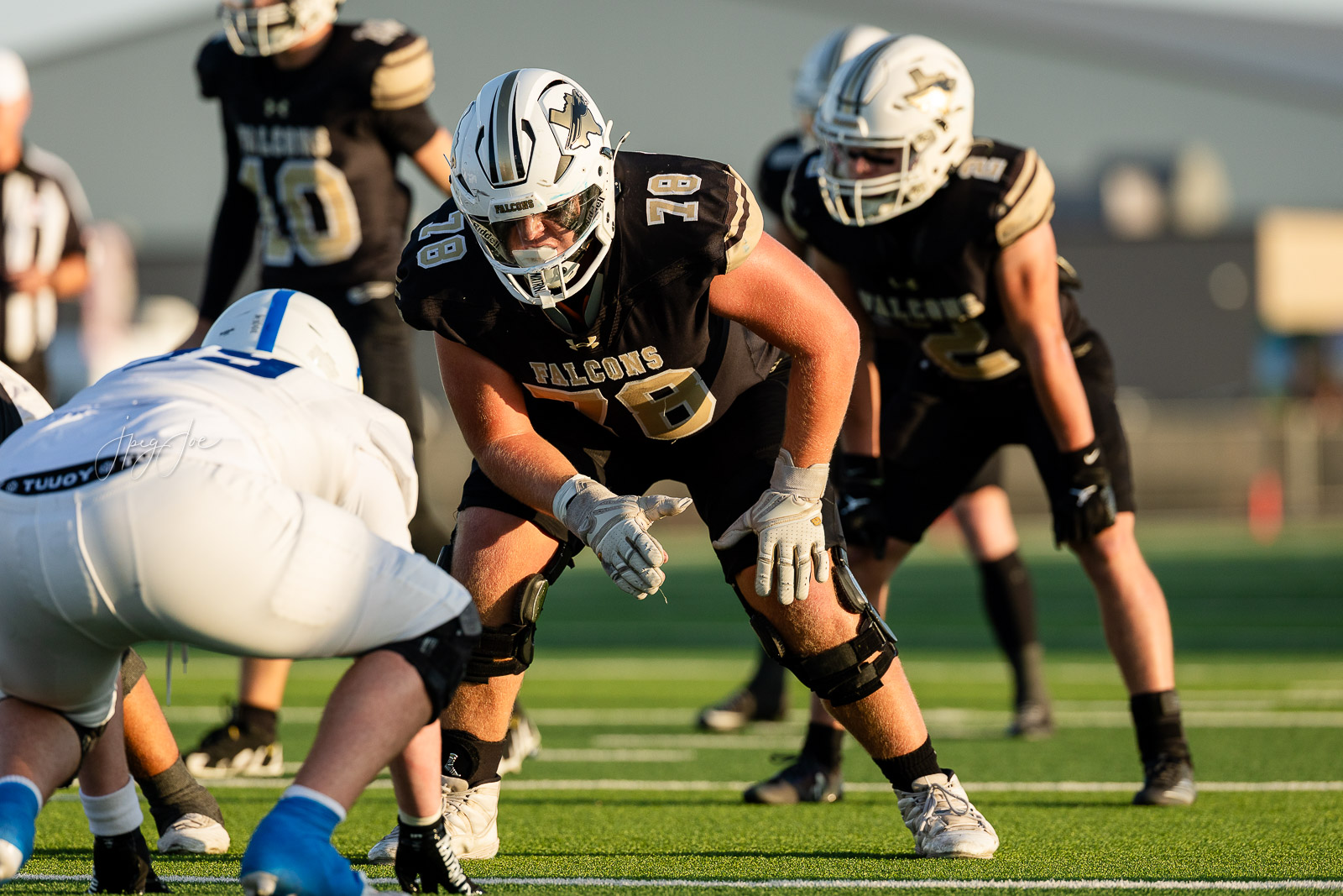 lubbock-christian-vs-bushland-football-jpegJoe-10 | Press Pass Sports Falcons offensive lineman, number 78, crouches at the line of scrimmage ready to snap the ball in a black and gold jersey. The opposing team watches nearby on a sunny field.