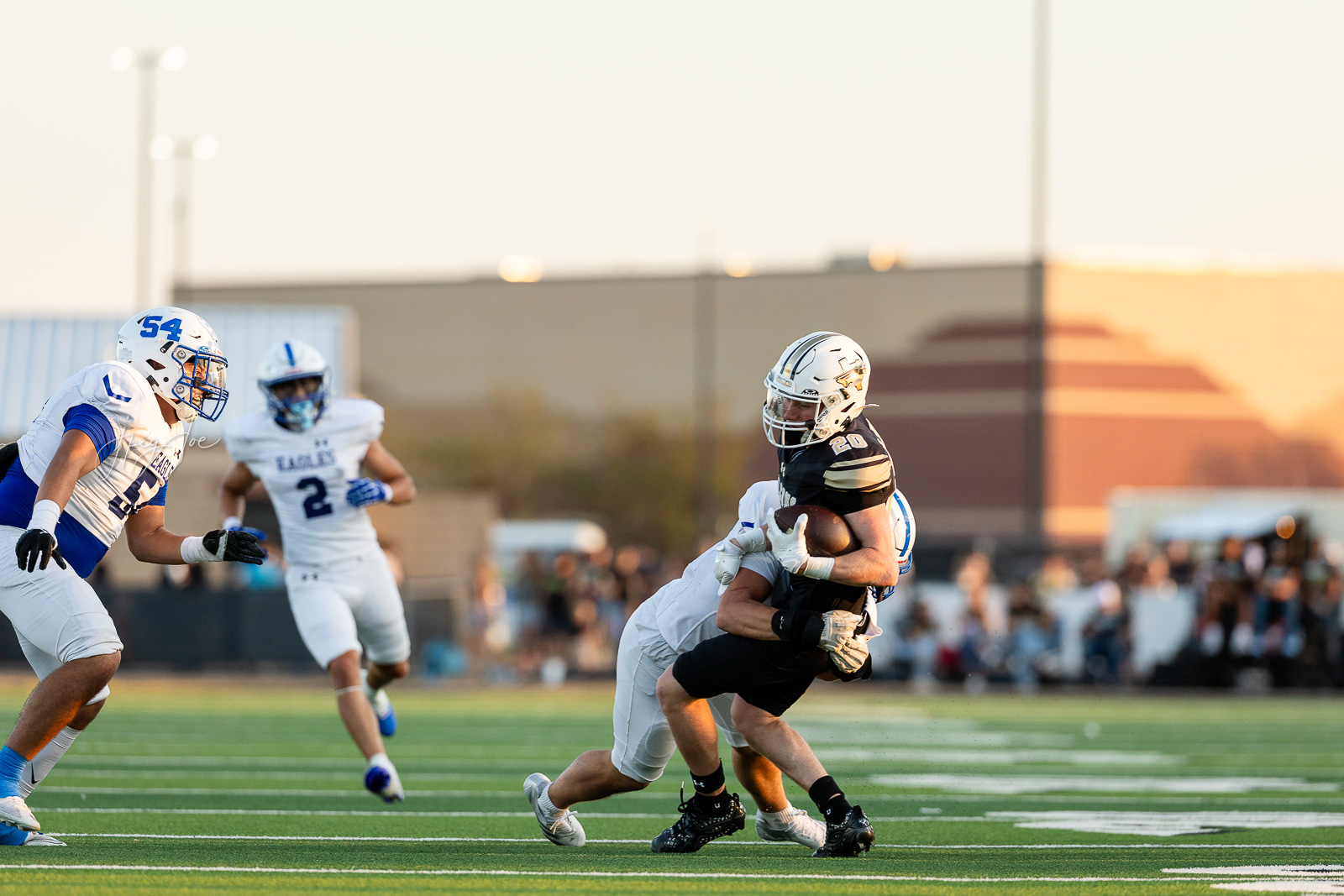 lubbock-christian-vs-bushland-football-jpegJoe-13 | Press Pass Sports Running back in black jersey with ball being tackled by a white-jersey defender as teammates pursue on a sunny football field.