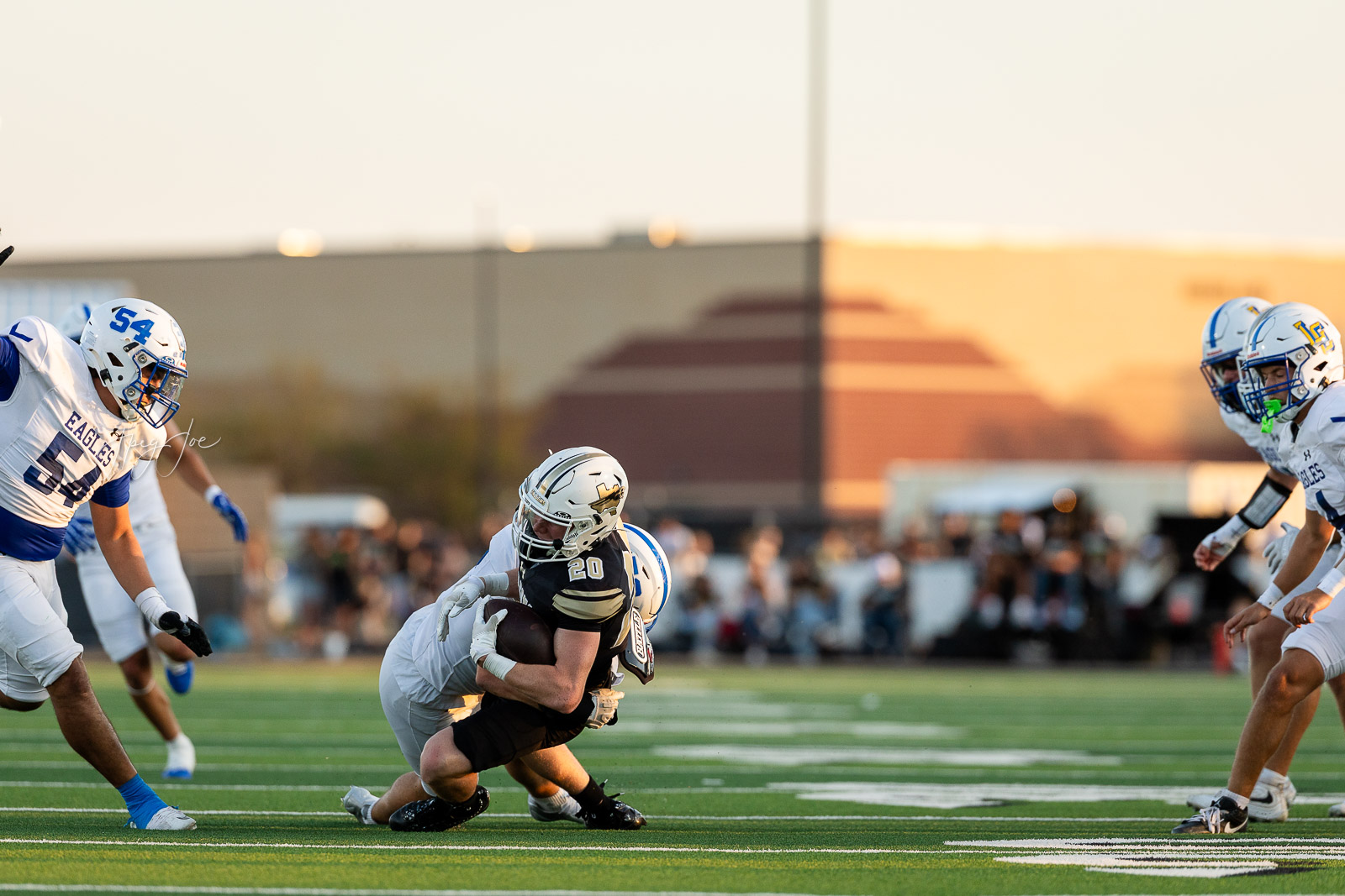 lubbock-christian-vs-bushland-football-jpegJoe-14 | Press Pass Sports Football player in black #20 is tackled by a defender on a sunlit field, with teammates and blurred crowd nearby.