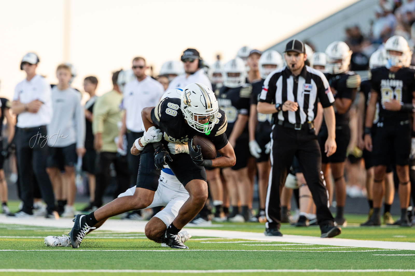 lubbock-christian-vs-bushland-football-jpegJoe-15 | Press Pass Sports Football player in black and gold jersey carries the ball while being tackled on a green turf field, with a referee nearby and teammates in the background.