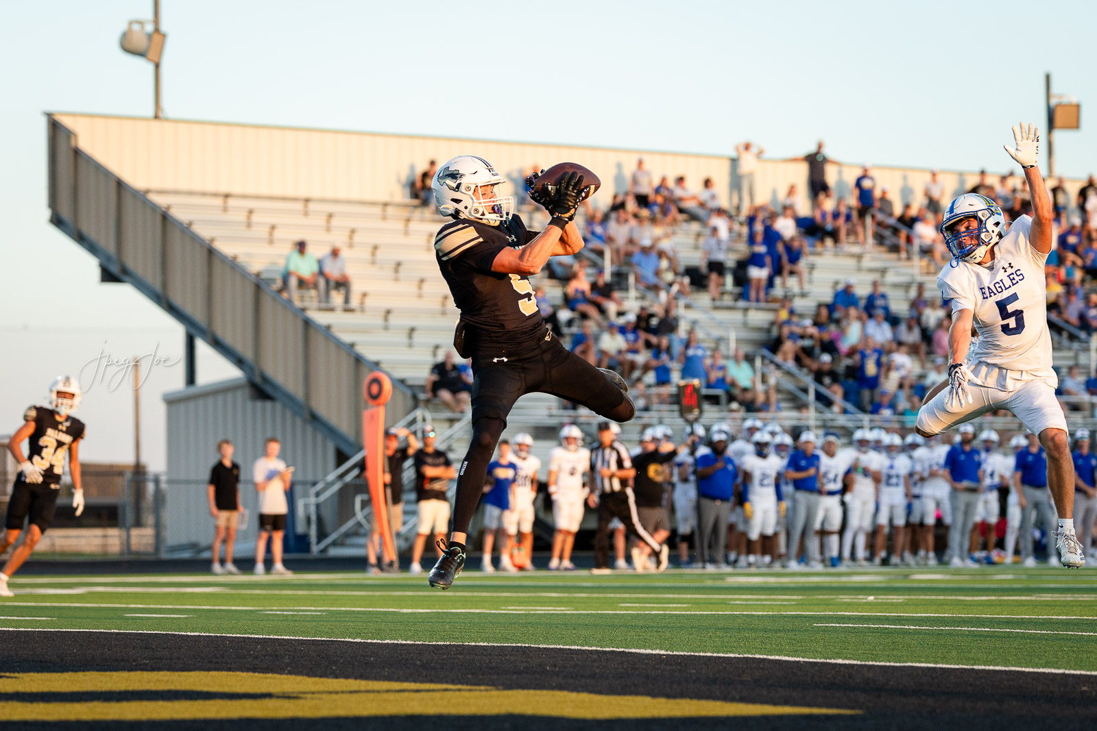 lubbock-christian-vs-bushland-football-jpegJoe-18 | Press Pass Sports Two football players jump for a caught pass on a sunlit field as a white-clad defender reaches in the background with spectators in the bleachers behind them.