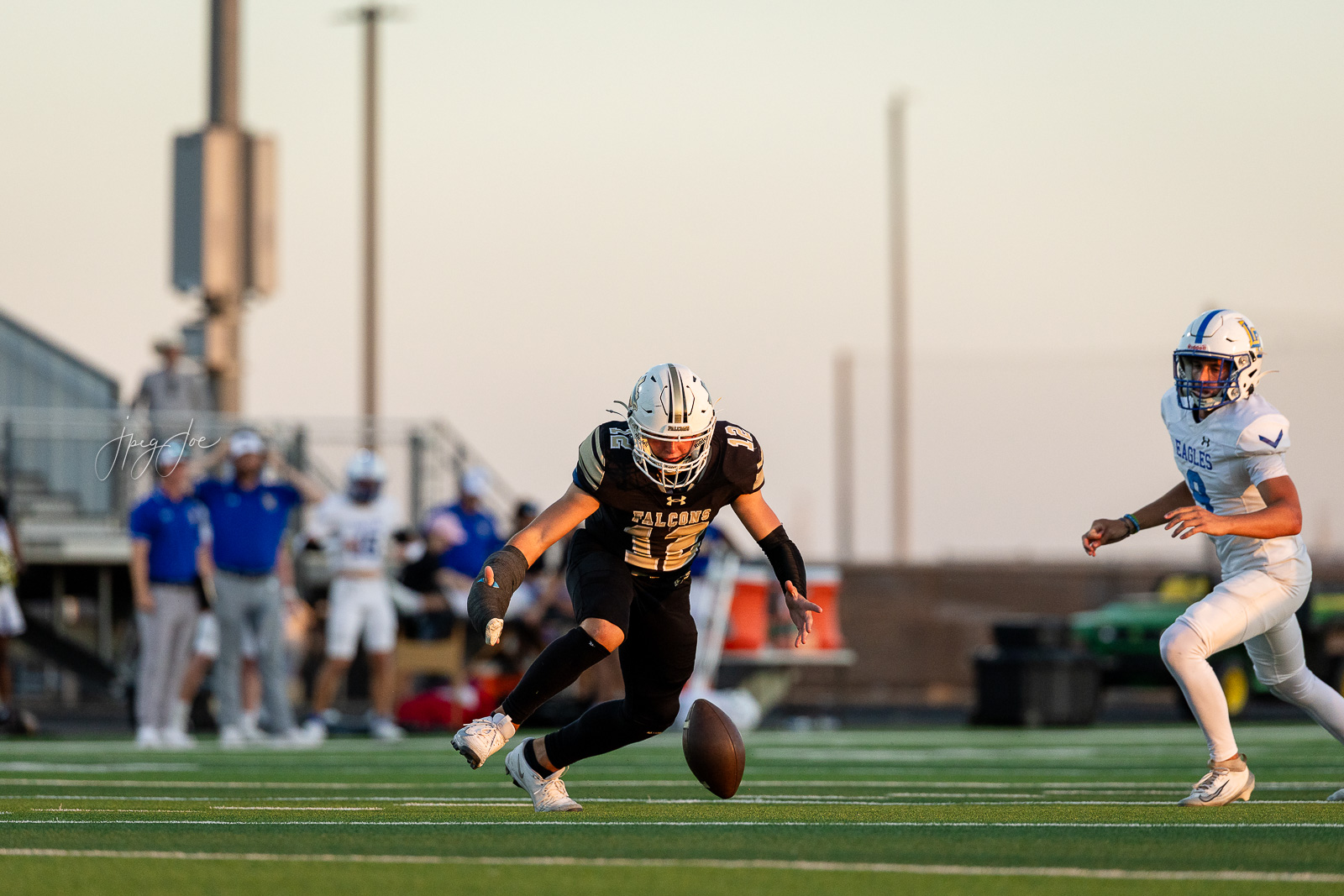 lubbock-christian-vs-bushland-football-jpegJoe-21 | Press Pass Sports A Falcons player in black jersey lunges for a loose football on a green field as an Eagles defender pursues from the right. Sunset light casts long shadows in the background.
