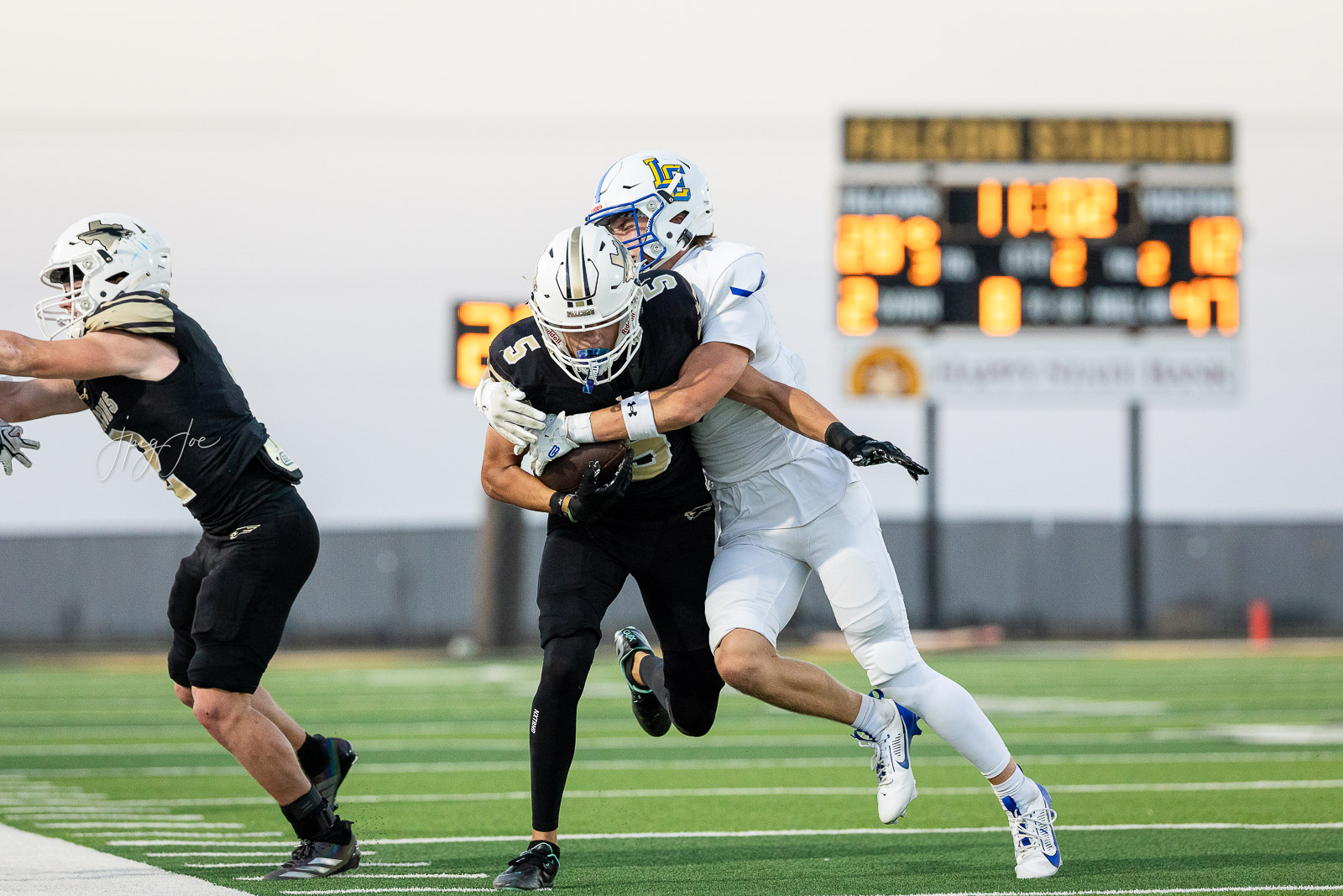 lubbock-christian-vs-bushland-football-jpegJoe-25 | Press Pass Sports Two football players collide as a ball carrier is tackled on a green turf field, with a scoreboard in the background.