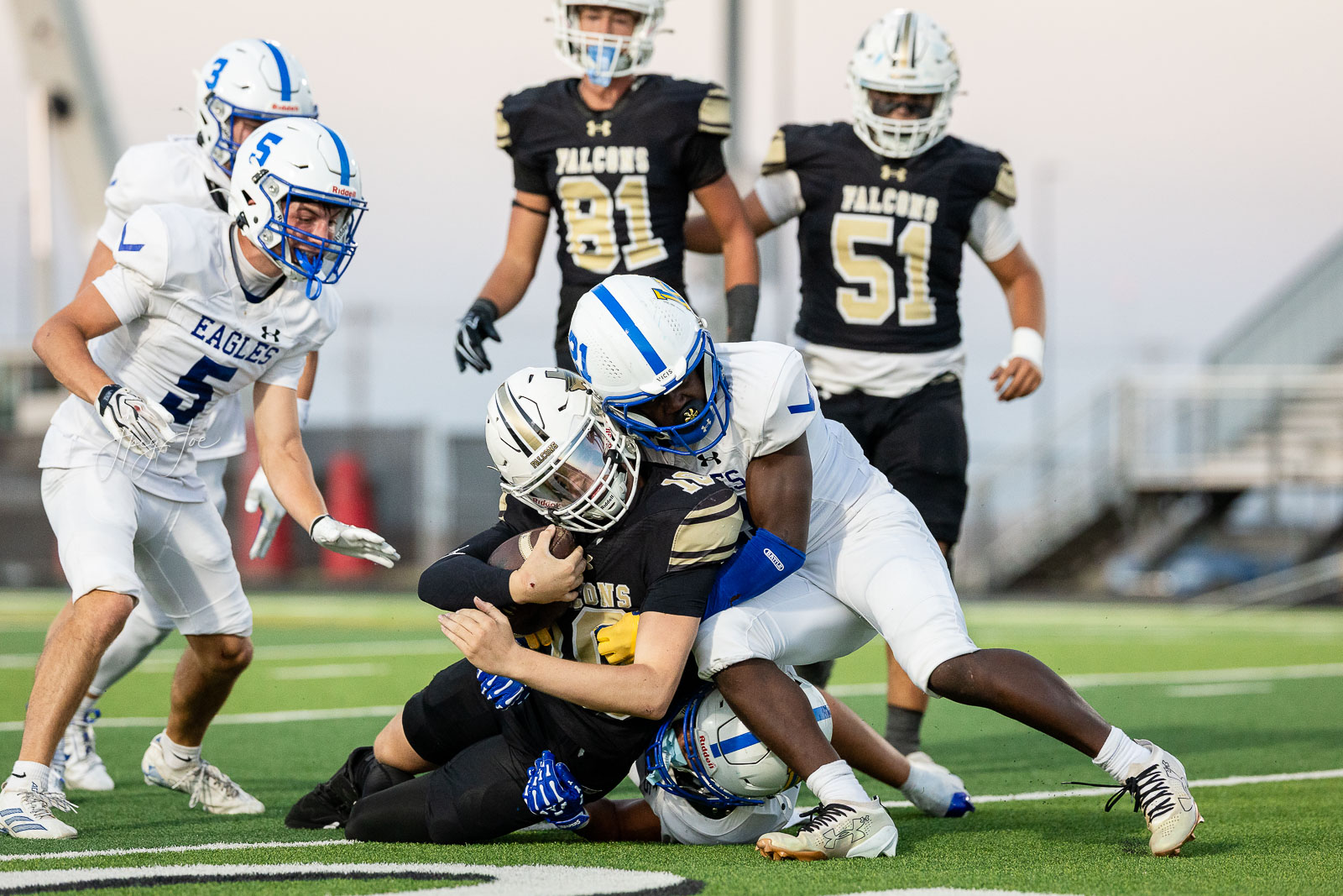 lubbock-christian-vs-bushland-football-jpegJoe-26 | Press Pass Sports White Eagles player wraps arms around a ballcarrier on a green turf during a high school football game as teammates close in nearby and defenders pursue fall in the background.