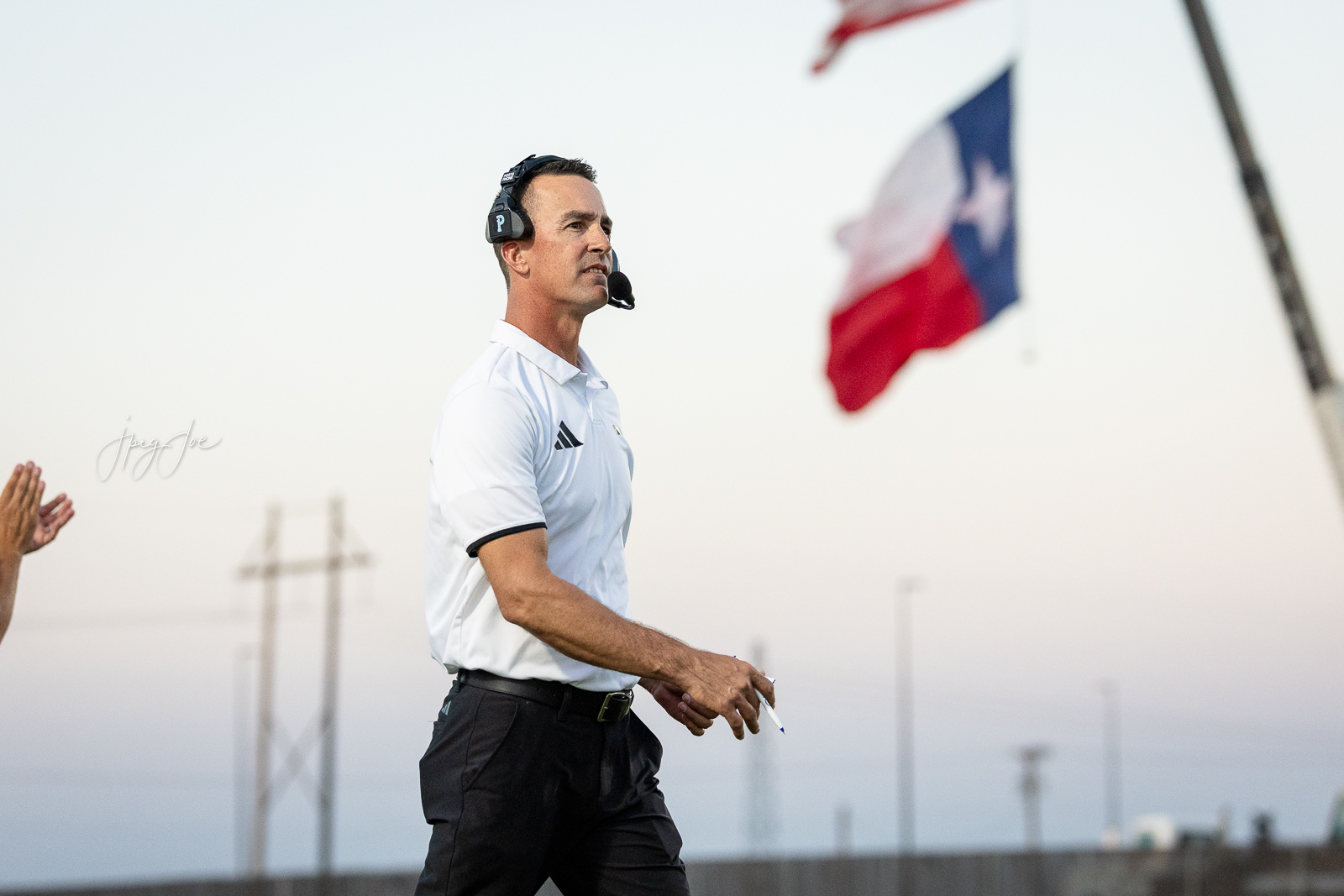 lubbock-christian-vs-bushland-football-jpegJoe-27 | Press Pass Sports Coach wearing a headset walks on a field, holding a pen with a flag in the blurred background