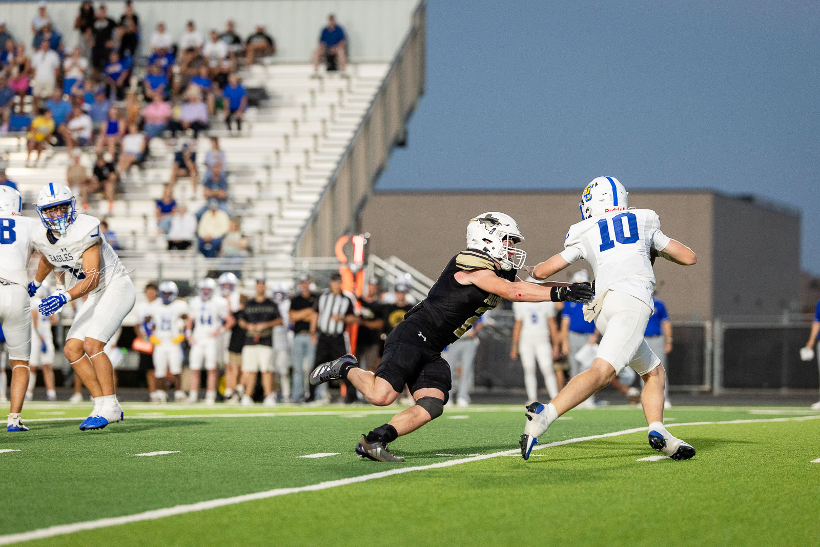 lubbock-christian-vs-bushland-football-jpegJoe-28 | Press Pass Sports Black-uniform defender tackles white-uniform player wearing #10 during a football play on a green field, with a crowd in the bleachers behind them.
