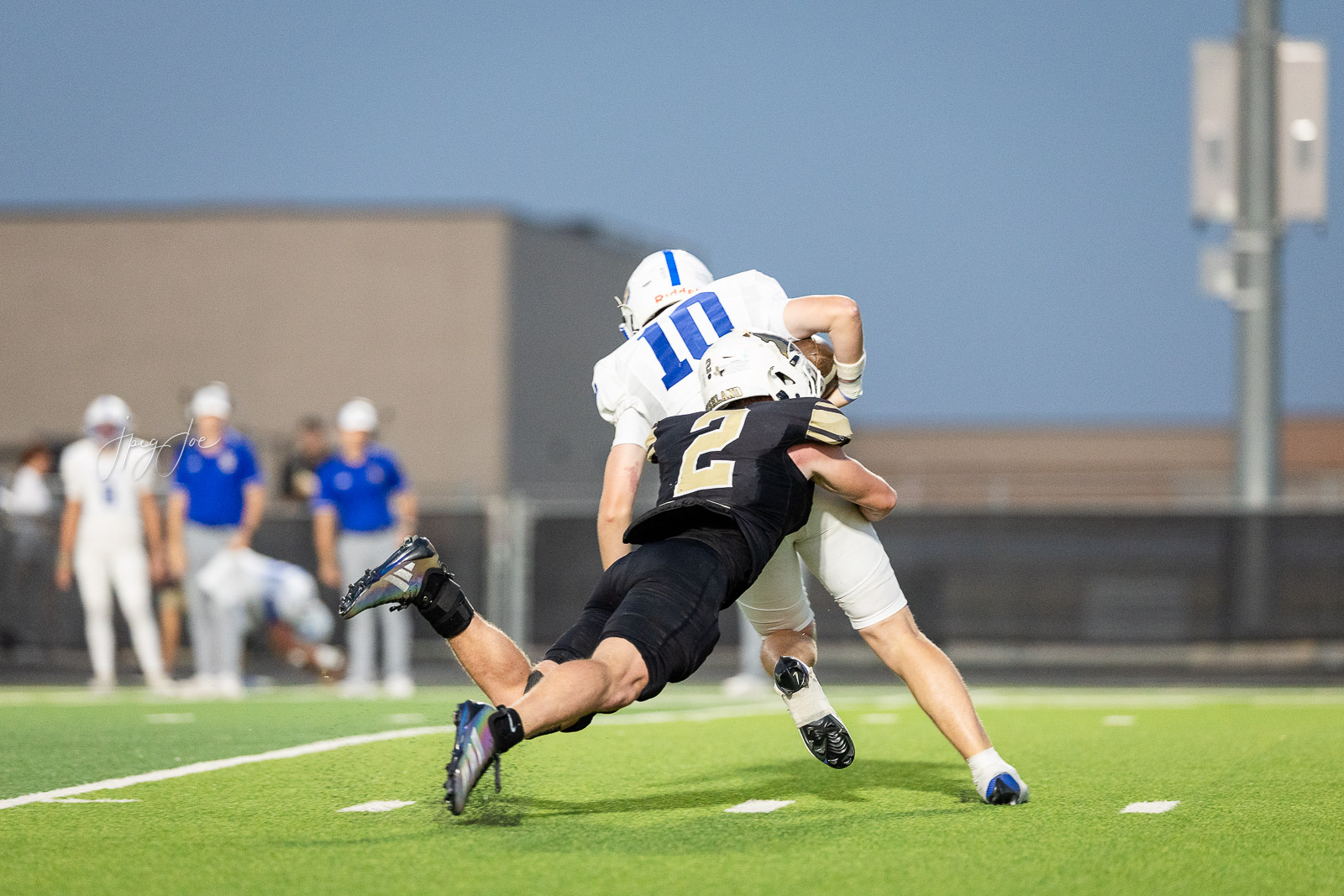 lubbock-christian-vs-bushland-football-jpegJoe-29 | Press Pass Sports Two football players collide in a tackle on a bright green turf; white jersey with blue #10 tackled by a black jersey with gold #2