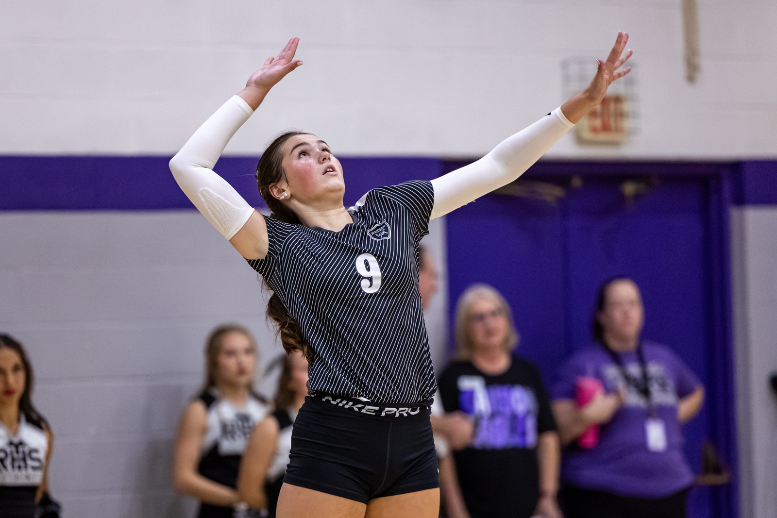 CHS-RHS-VB-4 1 | Press Pass Sports Female volleyball player in a striped jersey with number 9 raises arms in a jump, preparing a spike in a gym.