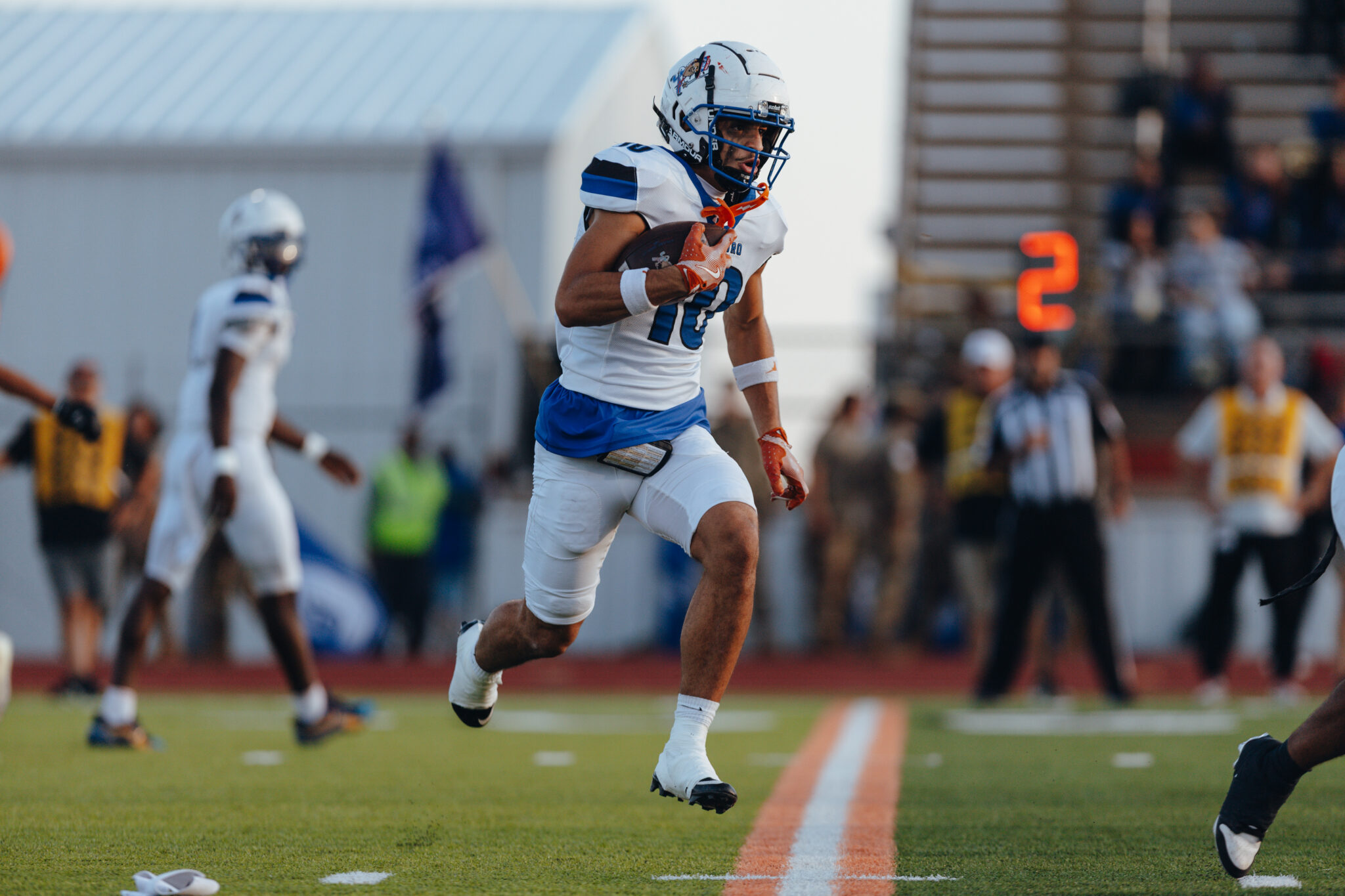 2C5A2549 | Press Pass Sports Football player in white and blue uniform running with the ball on a turf field, with a blurred crowd in the background.