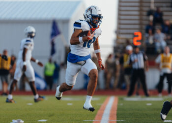 2C5A2549 | Press Pass Sports Football player in white and blue uniform running with the ball on a turf field, with a blurred crowd in the background.