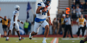 Football player in white and blue uniform running with the ball on a turf field, with a blurred crowd in the background.