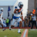 Football player in white and blue uniform running with the ball on a turf field, with a blurred crowd in the background.