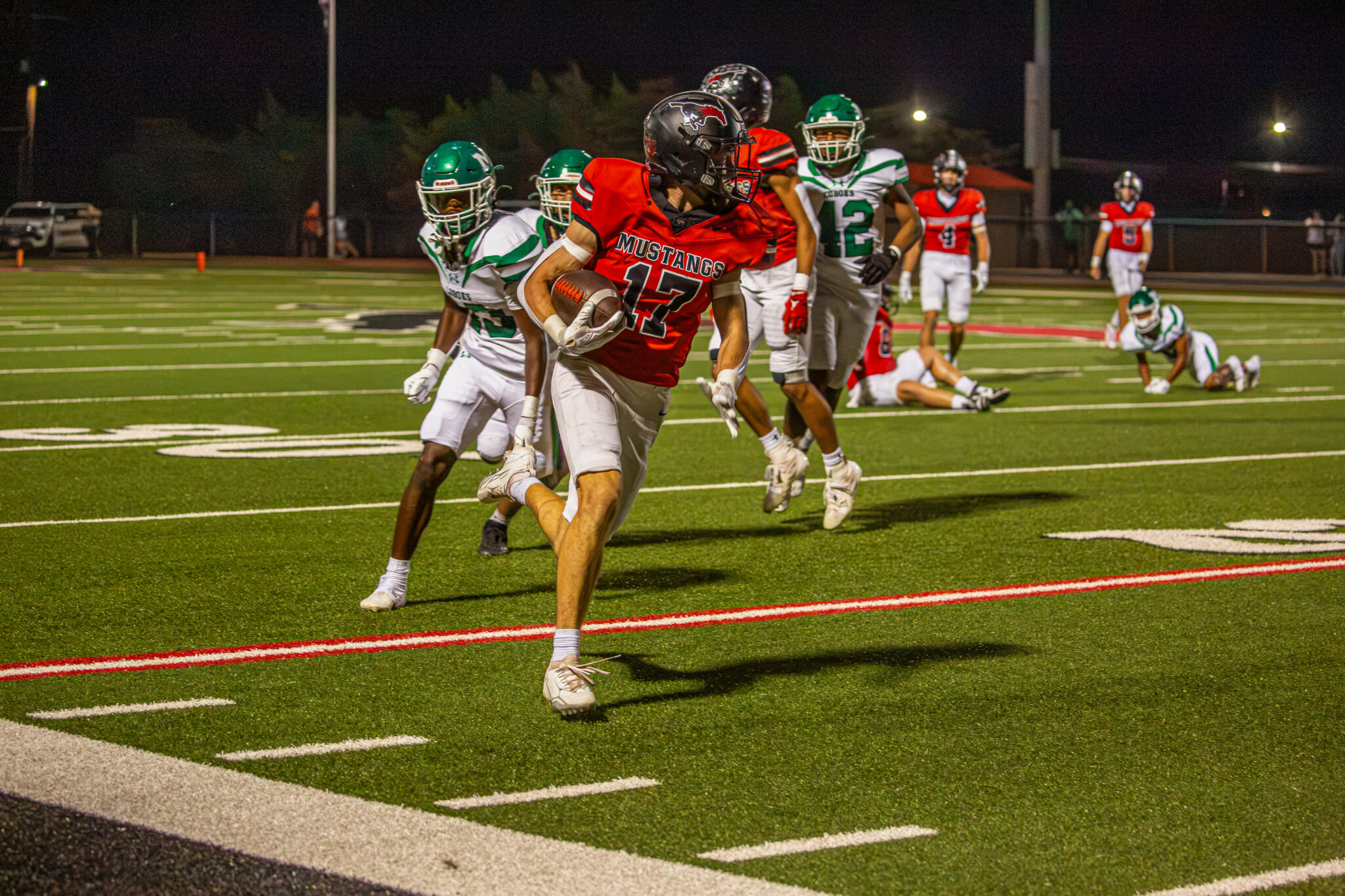 _V4A4469 | Press Pass Sports Rushing Mustang #17 carries the football while defenders in green pursue during a night game on an artificial turf field.