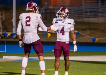 Simmons ASU | Press Pass Sports Two football teammates in white maroon uniforms give a high-five on a green field with bleachers in the background, celebrating.