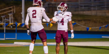 Two football teammates in white maroon uniforms give a high-five on a green field with bleachers in the background, celebrating.
