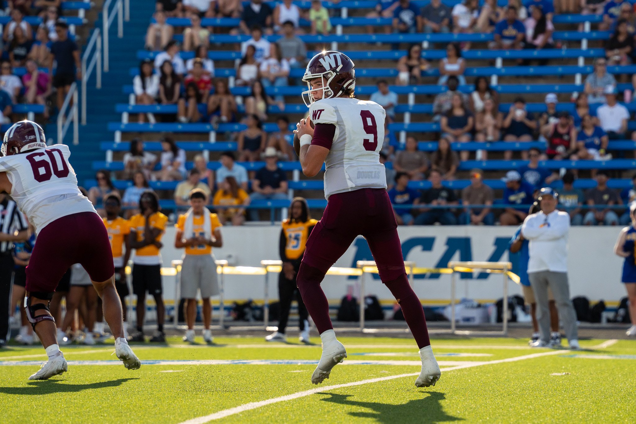 Quarterback #9 in white and maroon prepares to throw a pass as fans watch from blue bleachers in a daytime football game.