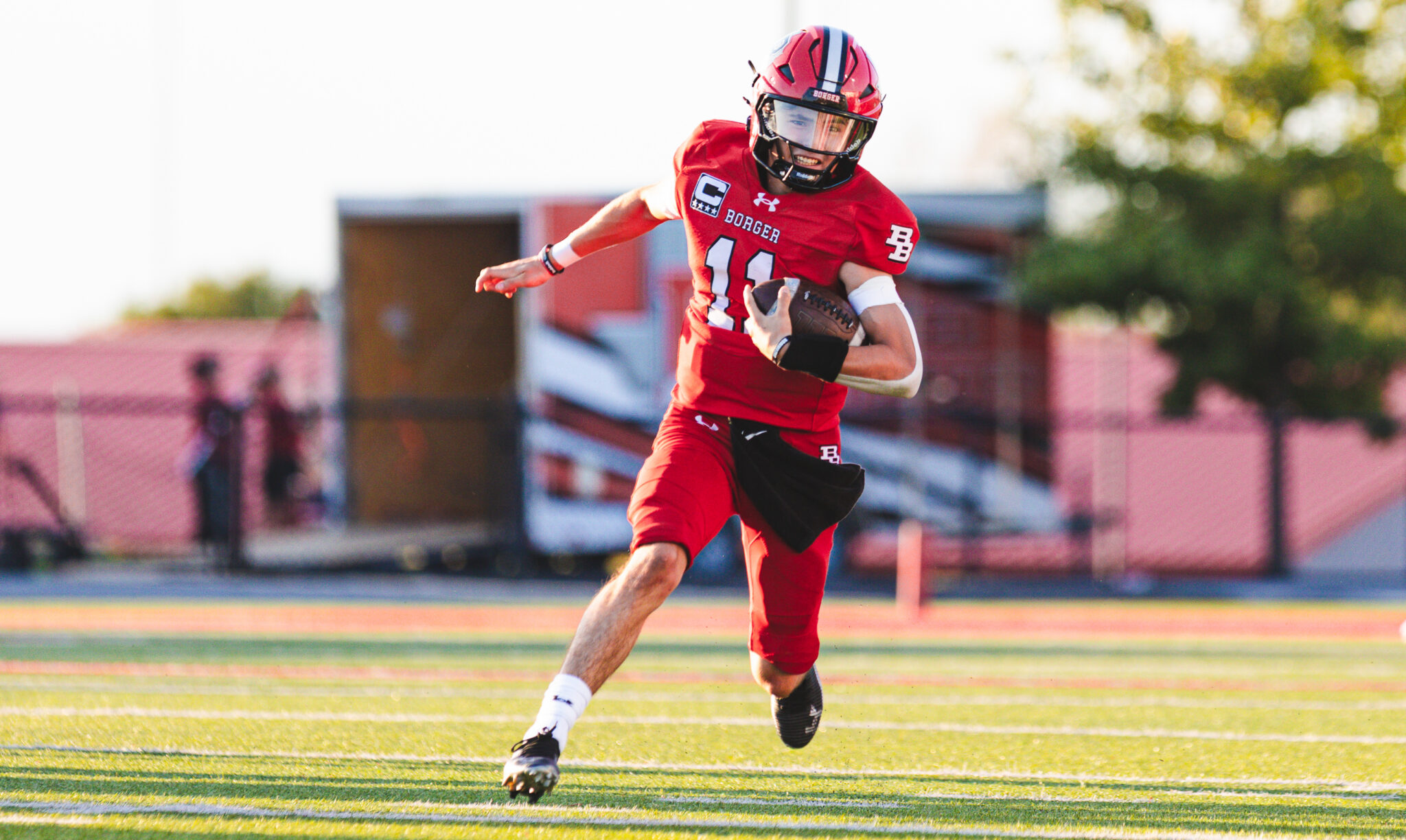 1H4A9529 | Press Pass Sports Football player in red uniform sprinting with ball during a game on a grassy field, wearing a red helmet and white wristbands.