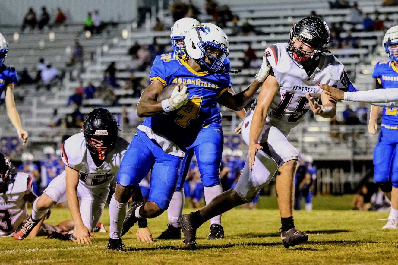 Blue team running back carries the ball while defenders reach in during a night football game on a lit field in a stadium.