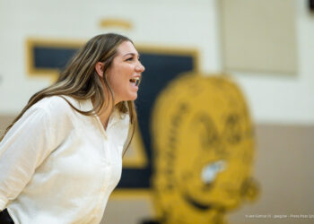 IMG_7524 | Press Pass Sports Woman in a white shirt laughing or shouting in a gym setting, with a large yellow mascot logo blurred in the background.