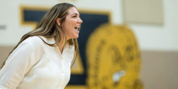 Woman in a white shirt laughing or shouting in a gym setting, with a large yellow mascot logo blurred in the background.