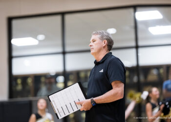 dumas-vs-bushland-volleyball-jpegJoe-pps-31 | Press Pass Sports Man in a black polo holding a clipboard with sheet music, standing in a gym while band members rehearse in the background.