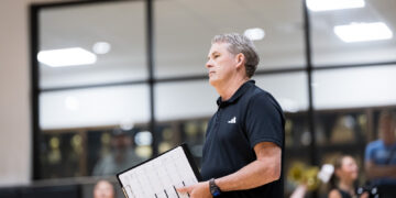Man in a black polo holding a clipboard with sheet music, standing in a gym while band members rehearse in the background.