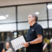 Man in a black polo holding a clipboard with sheet music, standing in a gym while band members rehearse in the background.