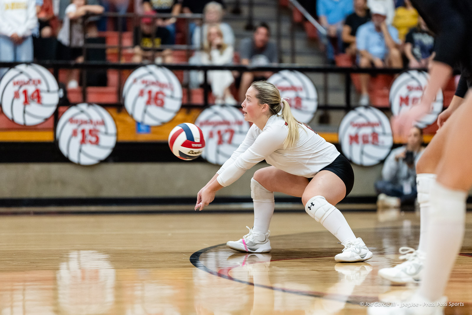amarillo-high-vs-tascosa-volleyball-jpegJoe-pps-2 | Press Pass Sports Female volleyball player in a white jersey and knee pads crouches to receive a ball during a match in a gymnasium with spectators in the background.