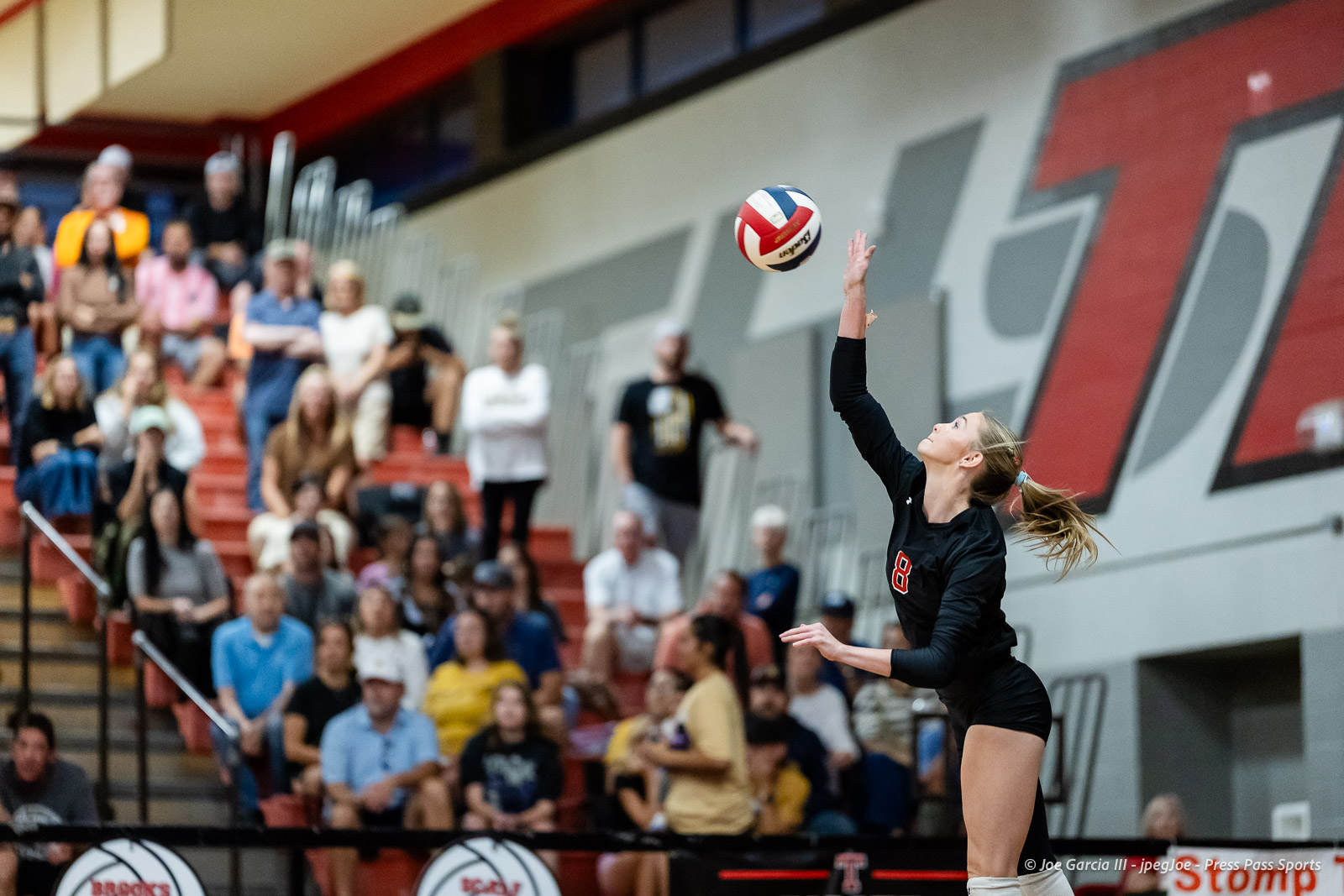 amarillo-high-vs-tascosa-volleyball-jpegJoe-pps-3 | Press Pass Sports Female volleyball player in a black uniform (number 8) serves the ball on an indoor court as spectators watch from red bleachers.