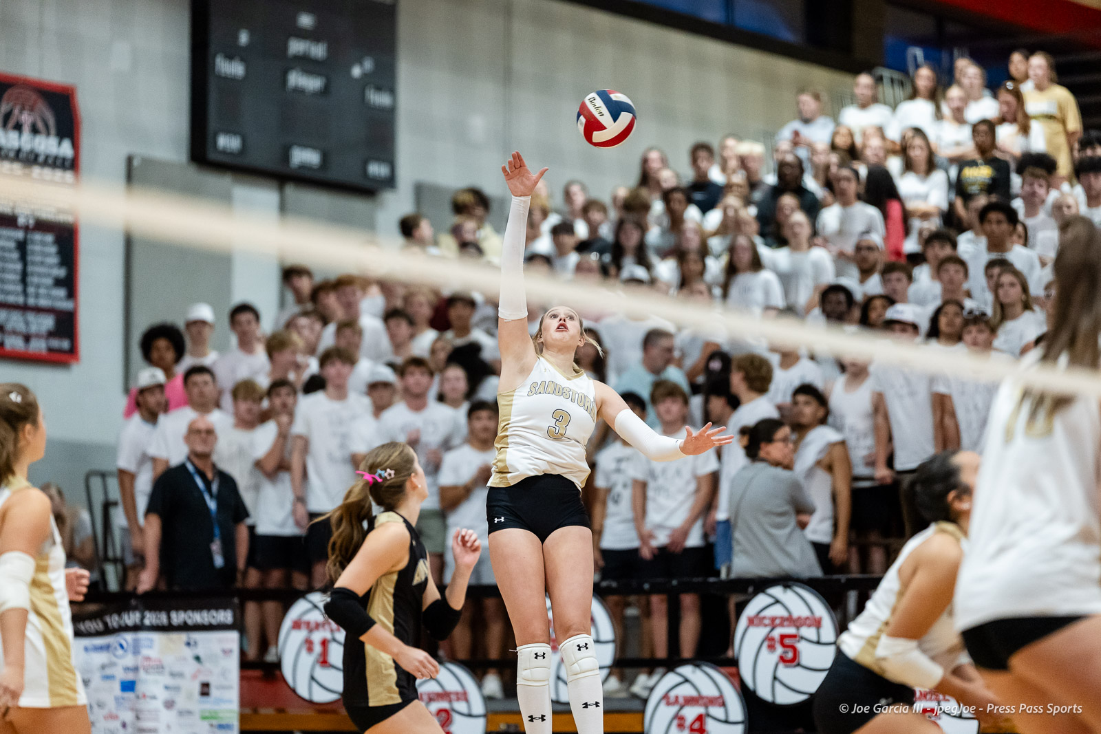amarillo-high-vs-tascosa-volleyball-jpegJoe-pps-5 | Press Pass Sports Female volleyball player in white and gold uniform leaps to spike the ball as a large crowd watches from the stands in a gymnasium.