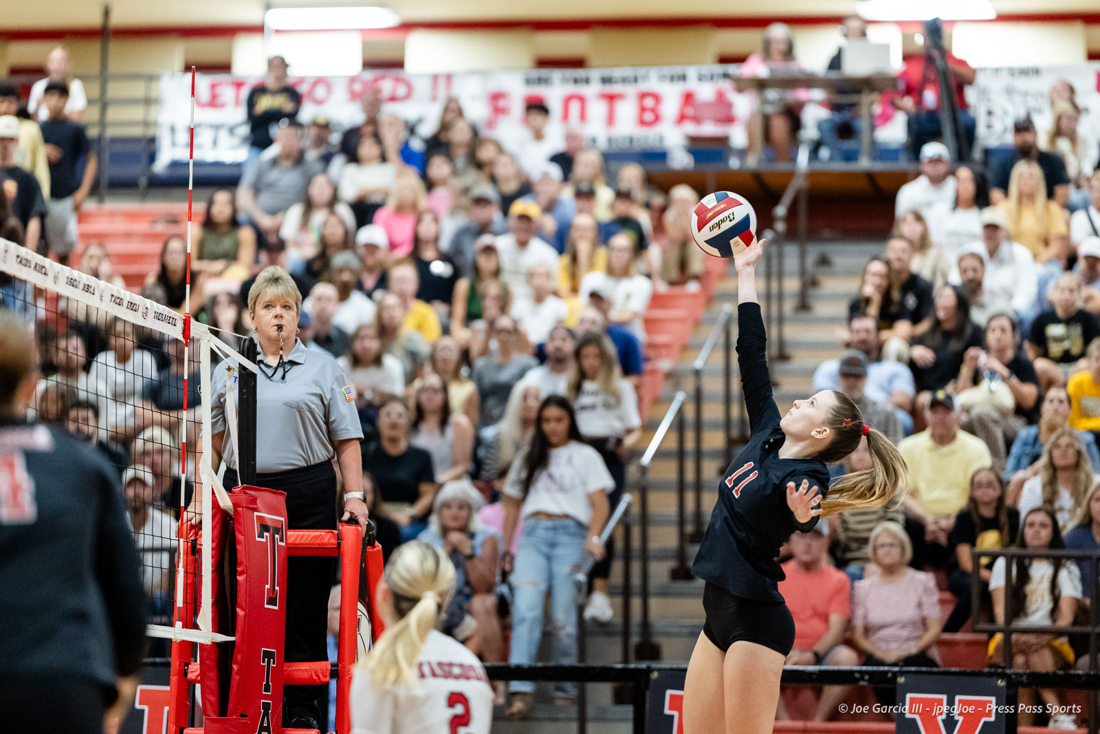 amarillo-high-vs-tascosa-volleyball-jpegJoe-pps-7 | Press Pass Sports Female volleyball player in a black uniform reaches to hit the ball over the net as the referee watches, with a crowded gym audience in the background in a sports event.Chair and banner visible in stands as fans observe the match (informative).