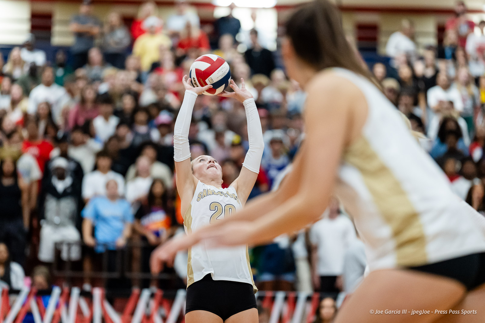 amarillo-high-vs-tascosa-volleyball-jpegJoe-pps-11 | Press Pass Sports Female volleyball player in white and gold jersey (#20) reaches up to set the red-white ball as a cheering crowd watches from the stands.