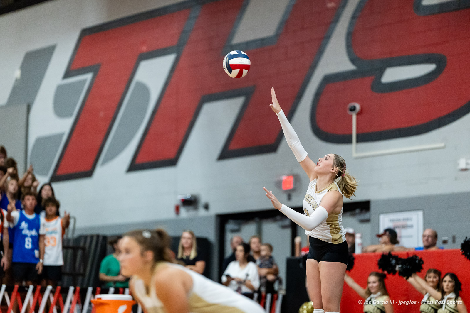 amarillo-high-vs-tascosa-volleyball-jpegJoe-pps-13 | Press Pass Sports Female volleyball player in gold/white uniform serves the ball during a match as crowd watches in a gym with large red/gray wall art behind her