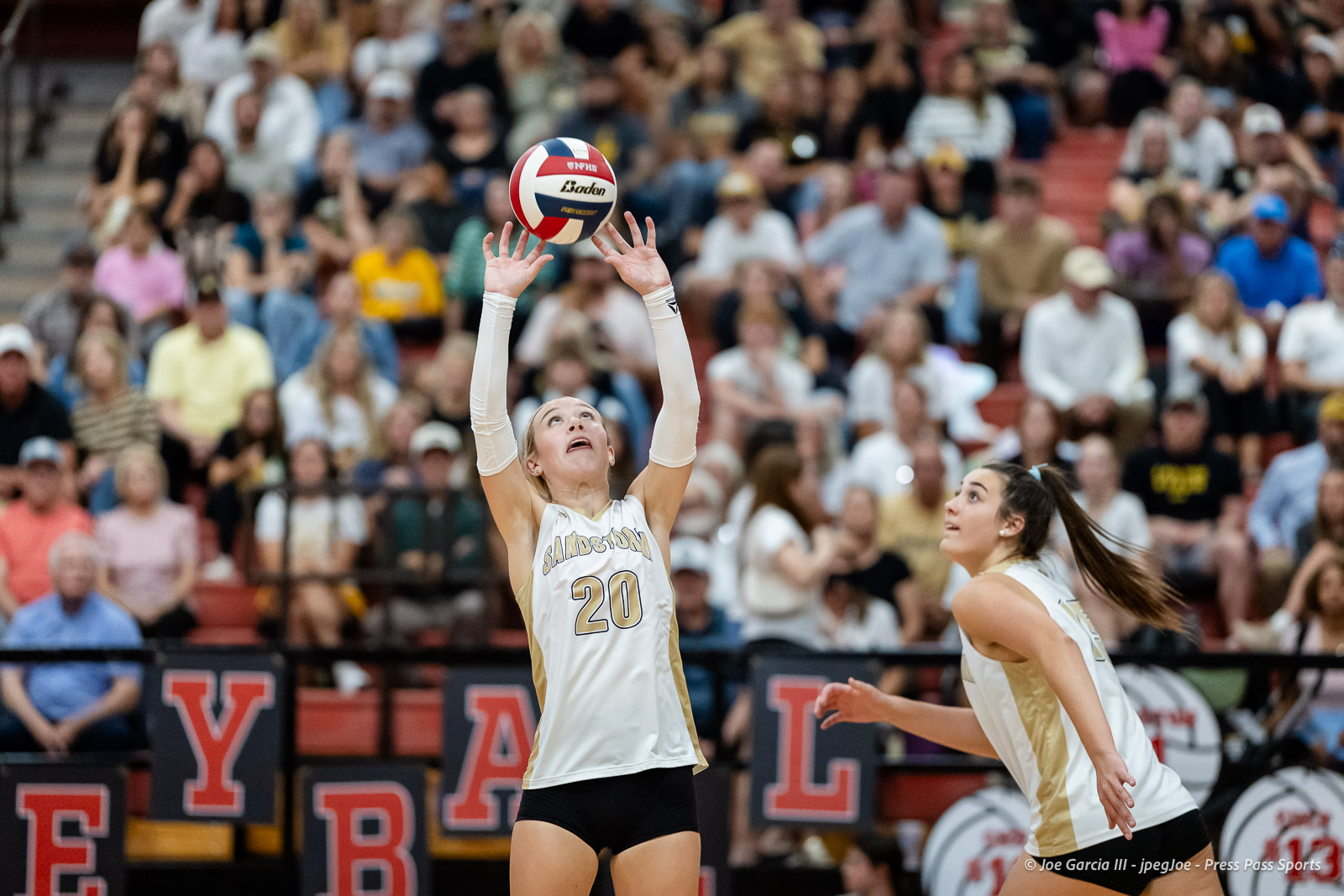 amarillo-high-vs-tascosa-volleyball-jpegJoe-pps-18 | Press Pass Sports Female volleyball player in white jersey number 20 jumps with arms raised to contact the ball overhead as a teammate watches, crowd in stands behind.