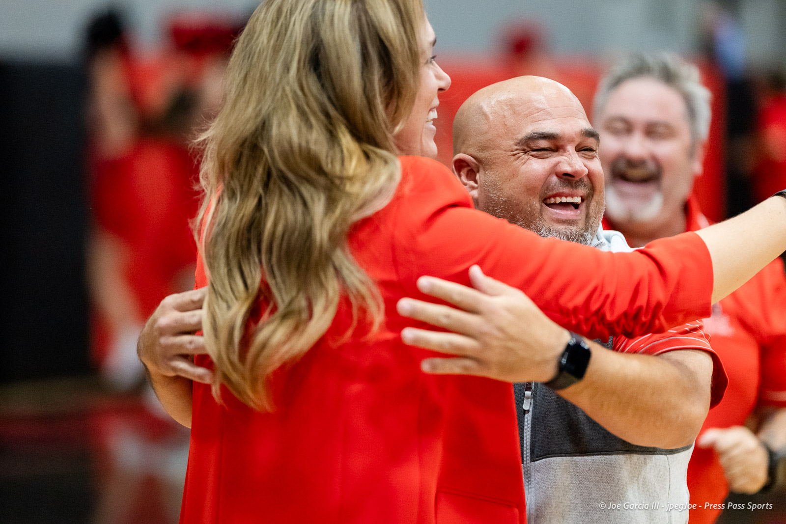 amarillo-high-vs-tascosa-volleyball-jpegJoe-pps-24 | Press Pass Sports A bald man and a woman with long blonde hair hug, smiling, surrounded by teammates in red uniforms at an indoor event in a gymnasium.